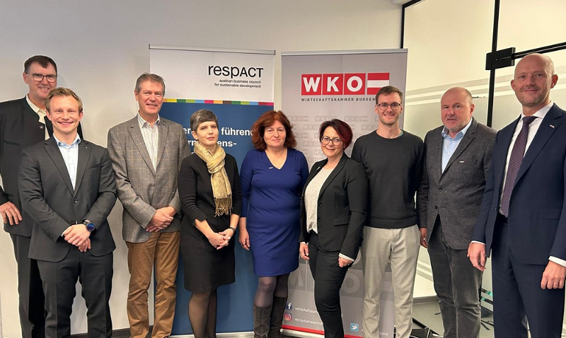 Eine Gruppe von neun Personen steht lächelnd vor zwei Roll-Up-Bannern in einem Büro. Die Personen tragen formelle Kleidung und posieren für ein Gruppenfoto.